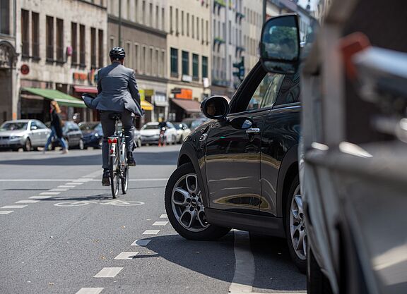Radfahrer in dunkler Kleidung mit Helm fährt auf einer Stadtstraße, im Vordergrund steht ein schwarzes Auto am Straßenrand, weitere Fahrzeuge und Gebäude im Hintergrund.